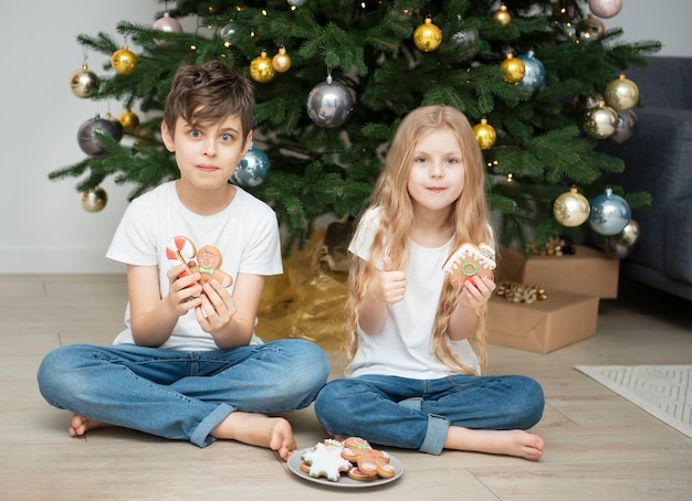 Children - a boy and a girl eating Christmas gingerbread near the Christmas tree in the living room