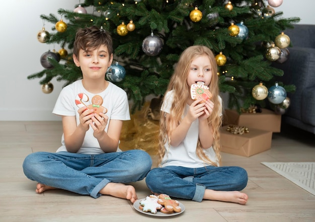 Children - a boy and a girl eating Christmas gingerbread near the Christmas tree in the living room