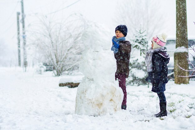 雪の降る冬の屋外の子供たちの男の子と女の子は大きな雪だるまを作っています