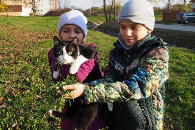 Children a boy and a girl of white race walk a black and white cat on the grassy lawn near the house The cat is afraid of a new place and tries to get out of hand Autumn sunny day