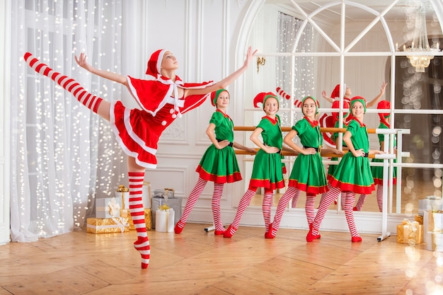 Children dressed as elves and practicing at a ballet barn look at a dancing ballerina dressed as santa claus in a spacious white studio.