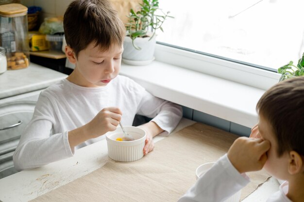子供たちは白いキッチンのテーブルで色付きのシリアルを食べます。 2人の男の子のための朝食。学校前の朝食用シリアル