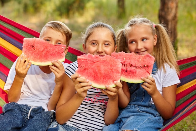 Children eat watermelon and joke outdoor sitting on a colorful hammock