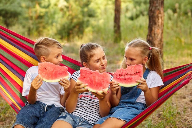 Children eat watermelon and joke outdoor sitting on a colorful hammock