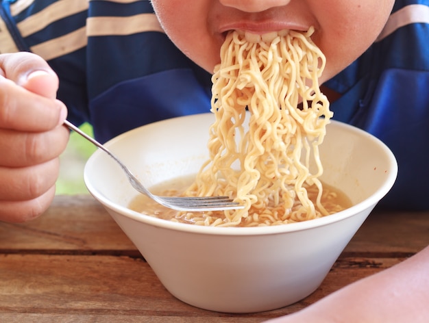 写真 木製のテーブルの白いボールでインスタントラーメンを食べる子供は、彼の口に焦点を当てました。