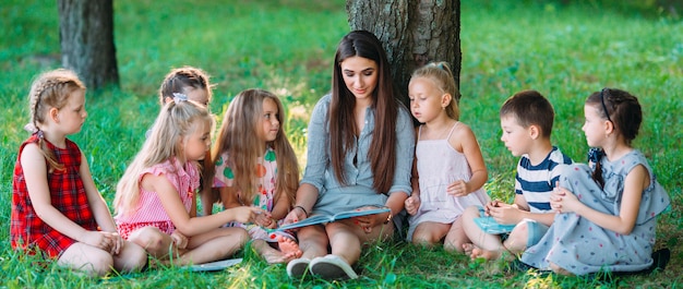 Bambini e istruzione, giovane donna al lavoro come educatore libro di lettura per ragazzi e ragazze nel parco.