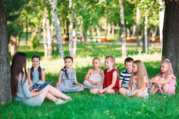 Foto bambini e istruzione, giovane donna al lavoro come educatore, leggendo un libro per ragazzi e ragazze nel parco