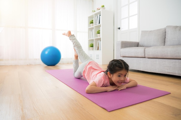 children girl doing yoga gym fitness action alone at home with big blue ball enjoying sporting stretching body lying on the floor in the room on sunny day afternoon.