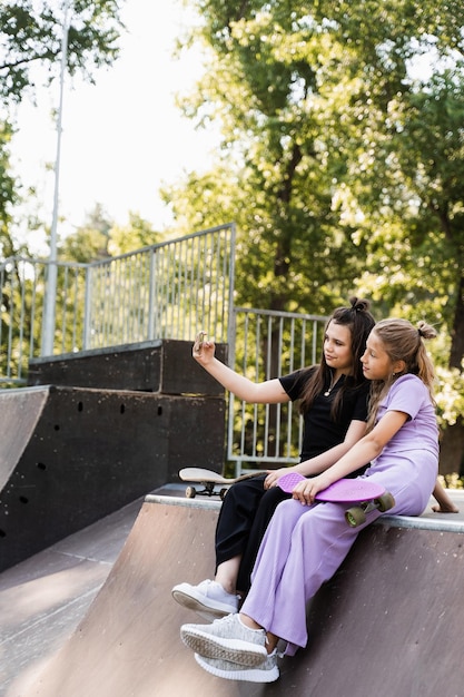 Children girls with skateboard and penny boards making selfie on phone on the sports playground Children friendship concept Kids friends smile and laugh and have fun together