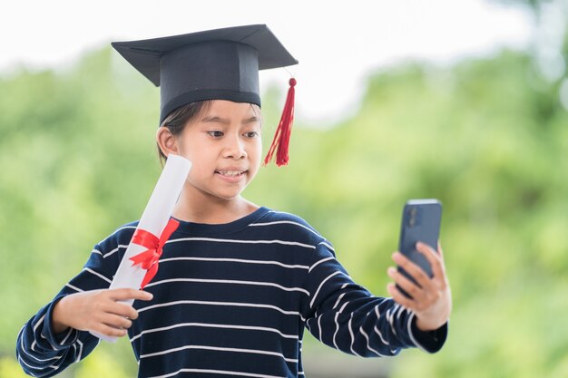 Children graduate with graduation cap and rolled graduation certificate holding a smartphone to call