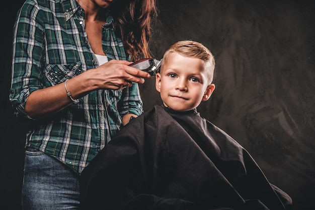 Children hairdresser with the trimmer is cutting little boy against a dark background. Cute preschooler boy getting haircut.