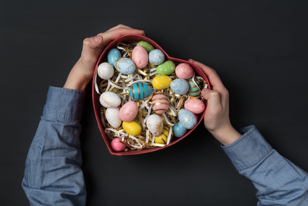 Children holding a Heart shaped box with colorful Easter eggs. Black background. Gift for Easter.