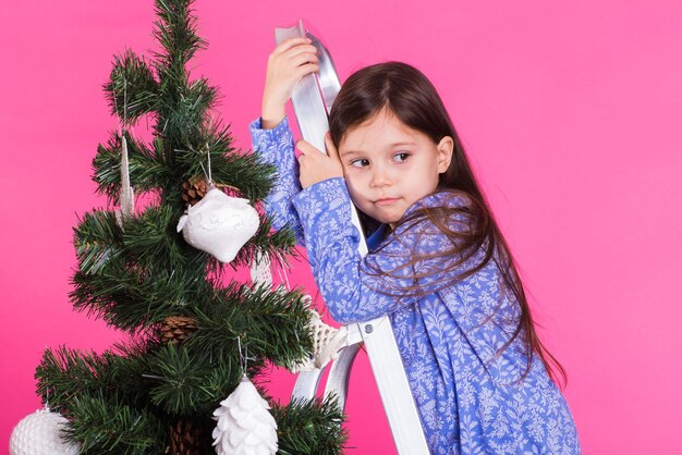 子供の休日とクリスマスのコンセプトピンクの背景にクリスマスツリーを飾る小さな女の子