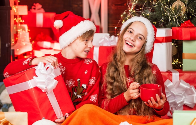 Children little happy girl and boy find gifts near christmas tree. Gifts shop. Kid santa hat christmas gift boxes. Merry christmas. Discover beauty of winter. Hot cocoa. Leisure and pleasant relax.