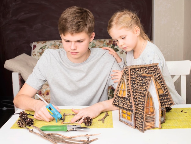 Children making house model of natural materials sticks and cones Teen boy and his  sister crafting