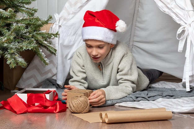 아이들은 크리스마스 선물을 엽니다 산타 모자를 쓴 십대 소년이 선물을 열고 바닥에 앉아 웃는다