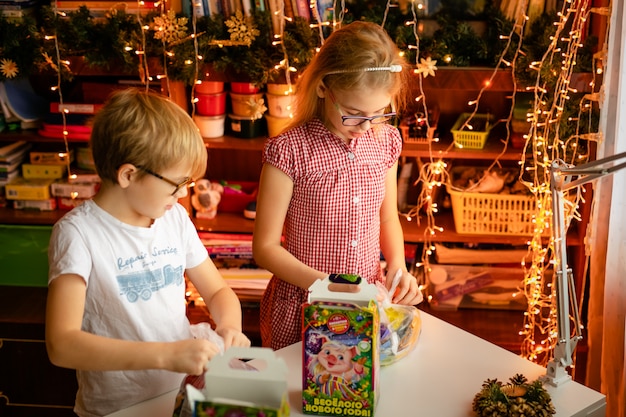 クリスマスプレゼントを開ける子供たち。小さな女の子とお菓子のプレゼントボックスを持つ少年。子供たちは贈り物を開きます。ギフト用の箱とキャンディーで遊ぶ子供たち