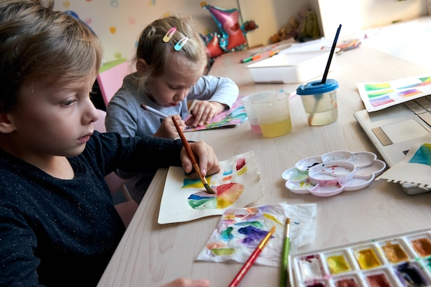 Children painting pictures with watercolor paints during art lesson. Pupils are concentrating on drawing with brush. Watercolor color wheel and palette. Color theory beginner hobby lessons