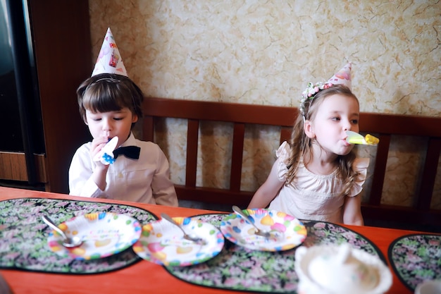 Children party in caps celebrating birthday with cake and balloons at home.