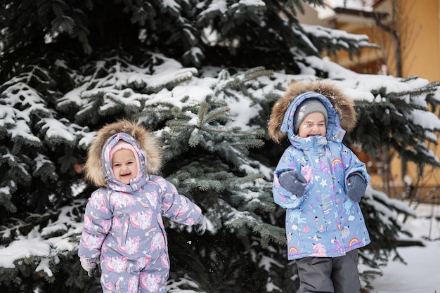 Children play outdoors in snow Two little sisters near Christmas tree in winter