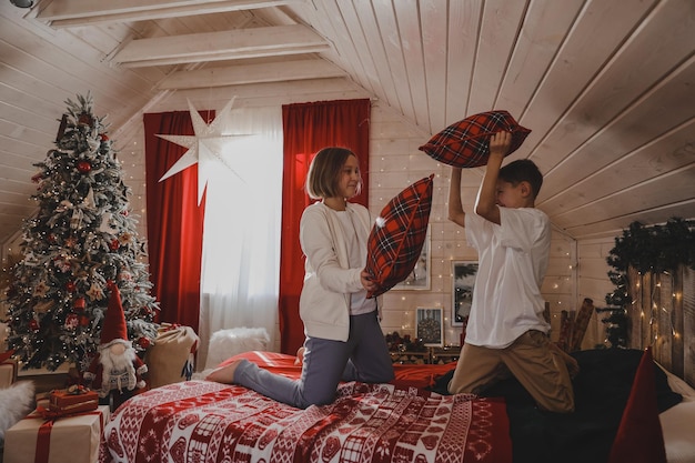 Children play with pillows on the bed in the Christmas-decorated bedroom, pillow fighting, the concept of the New Year holidays