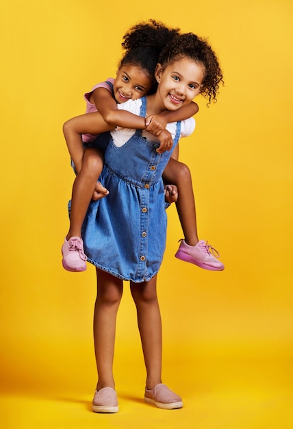 Photo children portrait and happy sisters for piggyback fun in studio with love of family on yellow background cute young girl kids or friends smile together for happiness play and support or care