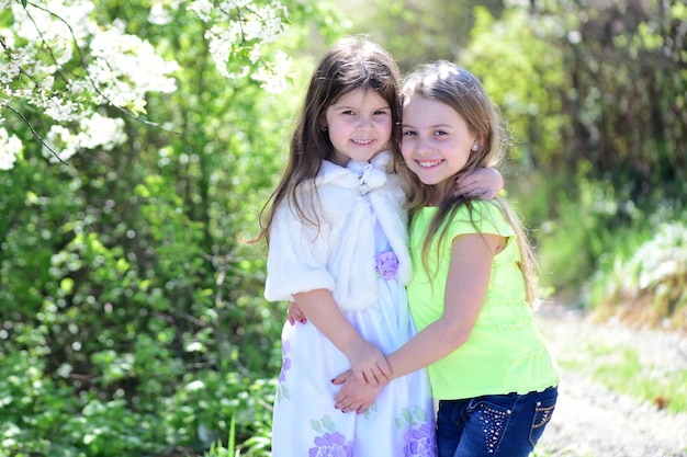 Children pose together on nature background defocused Sisters walk outside near white blooming apple trees Girls with smiling faces hug each other Childhood family and natural beauty concept