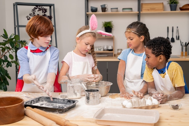 Photo children pouring dough together, wearing aprons looking at each other with wide smiles while preparing appetizing cookies for their parents