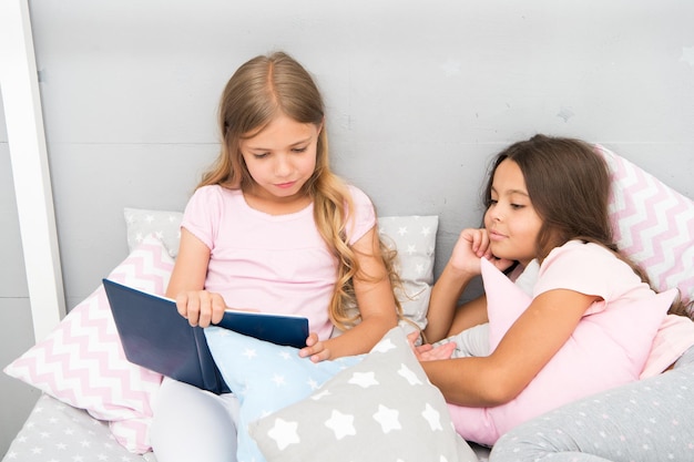 Children read book in bed. Stories every kid should read. Family tradition. Girls best friends read fairy tale before sleep. Best books for kids. Reading before bed can help sleep better at night.