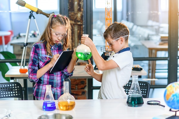 I bambini registrano i risultati degli esperimenti su notebook. due giovani allievi caucasici intelligenti in occhiali protettivi che fanno esperimento con liquido verde in becher e ghiaccio secco.