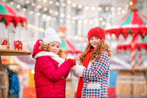 Children, red-haired sisters warm their gloved hands with a mug of hot tea at a festively decorated Christmas market in the city.