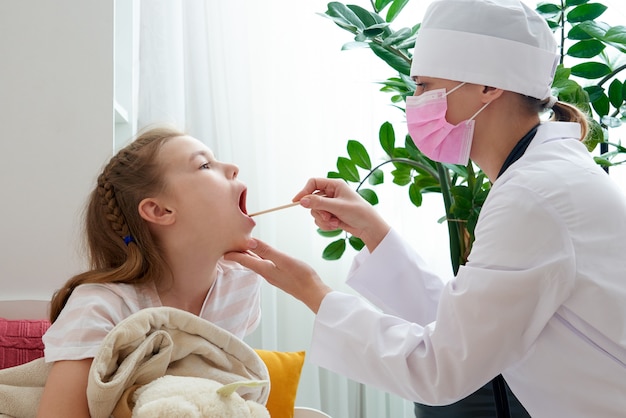 Фото Детский врач осматривает горло маленькой девочки дома с депрессором языка