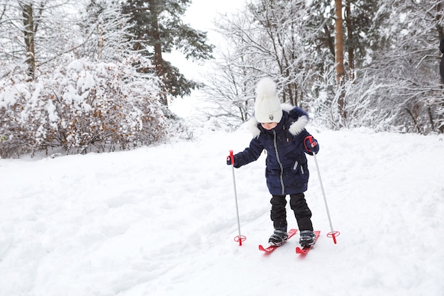 スティック付きの赤いプラスチックスキーの子供の足は、スライドから雪を通り抜けます-ウィンタースポーツ