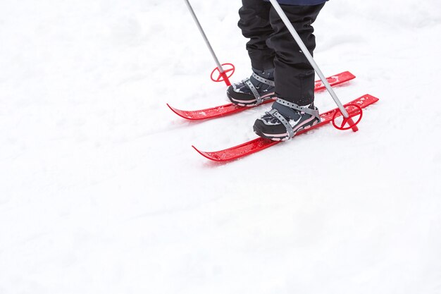 막대기가 달린 빨간색 플라스틱 스키를 타는 아이들의 발이 눈을 통과합니다.