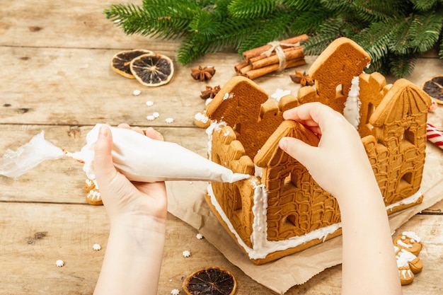 Children's hand decorate Gingerbread House by sweet icing. Handmade process, festive New Year decor. Traditional spices, wooden background, copy space