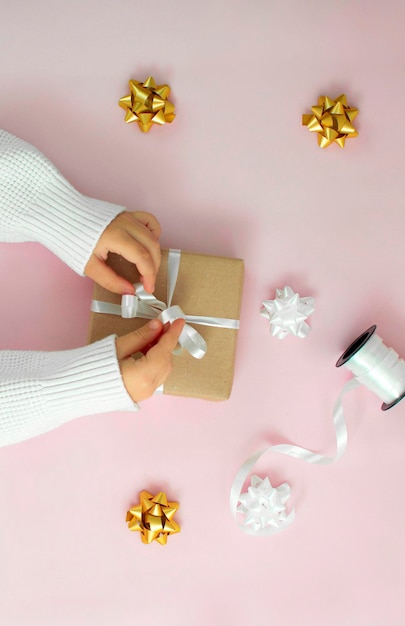 Le mani dei bambini tengono una confezione regalo kraft con un fiocco bianco su uno sfondo rosa. vista dall'alto, un concetto di regalo per le vacanze con spazio da copiare. stile di stile piatto