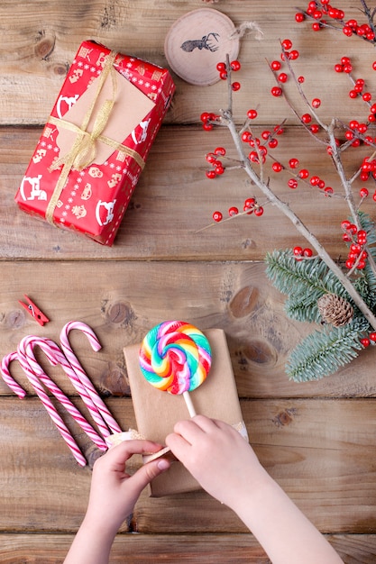 children's hands hold a lollipop with bright stripes and a gift on a wooden table and a branch with red berries