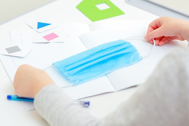 Children's hands holding a medical protective mask over school notebook on the desk at home. Quarantine studying concept.