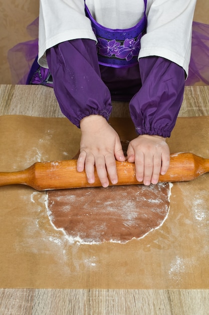 Children's hands roll out the dough with a rolling pin on a wooden table.