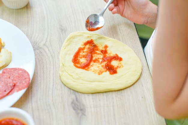 Children's hands with a spoon spreading ketchup on the dough Child little cook prepares pizza