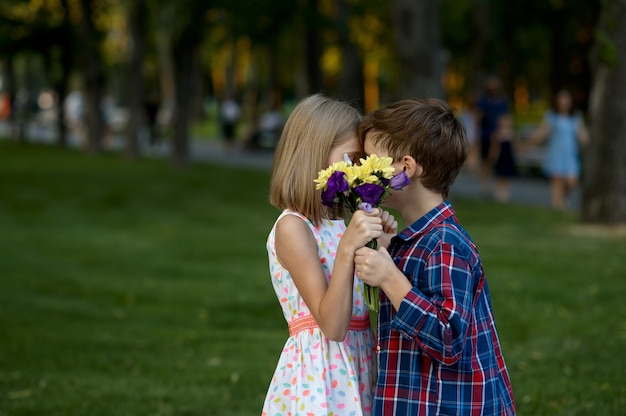 サマーパークでの子供のロマンチックなデート、友情、初恋のキス。散歩道に花束を持つ少年と少女。屋外で楽しんでいる子供たち、幸せな子供時代