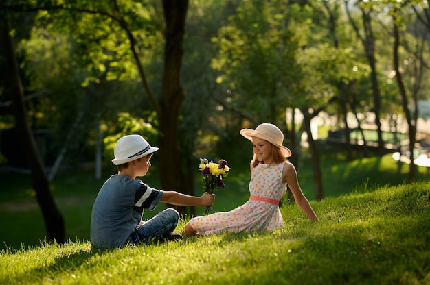 여름 공원에서의 아이들의 낭만적인 데이트, 우정, 첫사랑. 소년은 소녀에게 꽃다발을 줍니다. 야외에서 즐거운 시간을 보내는 아이들, 행복한 어린 시절