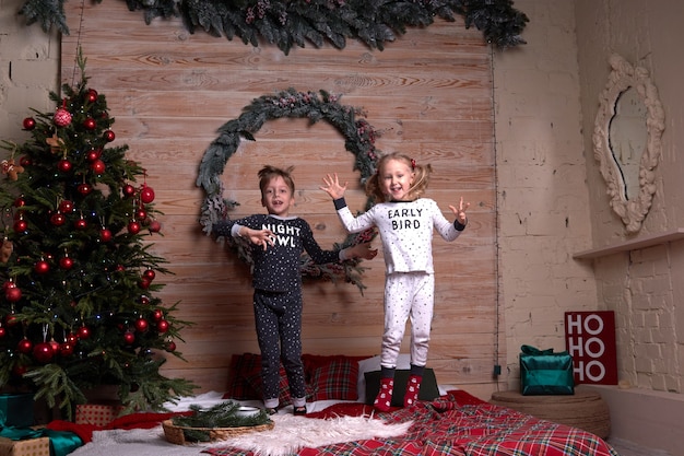 Children in the same family look soft warm pajamas playing at home in christmas evening under decorated Christmas tree. Happy kid jumping on the bed. New Year holidays.