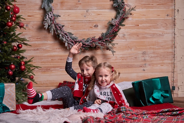 Children in the same family look soft warm pajamas playing at home in christmas evening under decorated Christmas tree. Happy kid jumping on the bed. New Year holidays.