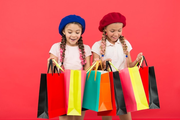 赤い背景の買い物に満足している子供たち。ショッピングや衣料品店に夢中。 Shopaholicのコンセプト。親友と一緒に買い物が楽しくなります。子供かわいい女子学生はたくさんの買い物袋を持っています。