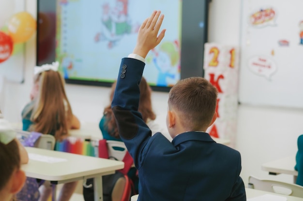 Children in school uniforms sitting by desks Boy raising hand to answer