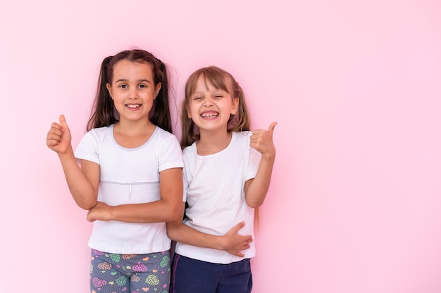 Children showing like. two adorable happy little girls\
gesturing thumbs up together and smiling to camera, excellent\
feedback, good job. indoor studio shot isolated on pink\
background