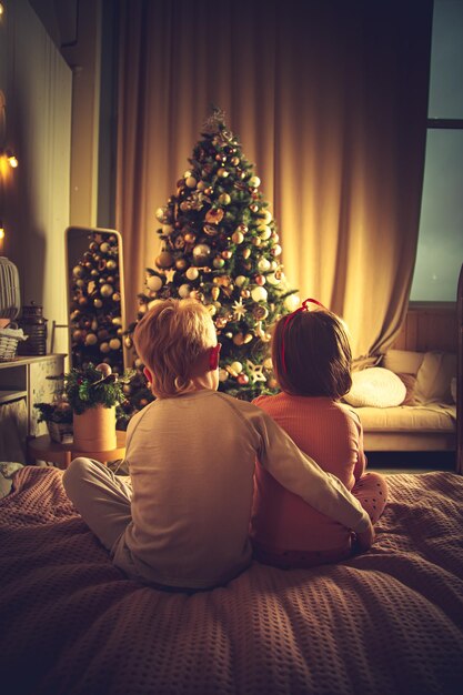 子供たちはベッドに抱きついて座って、クリスマスツリーを見ます。休日を待っているクリスマスの飾り。
