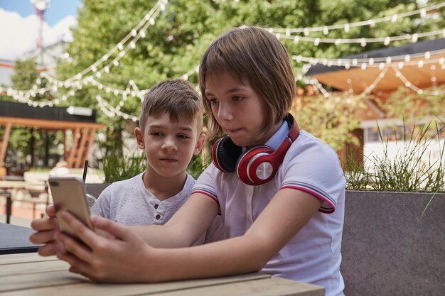 children sit outdoors, watch at screen of smartphone. little caucasian schoolchildren sitting at table at cafe veranda, using mobile phone. modern technology, gadgets, digital devices, entertainment