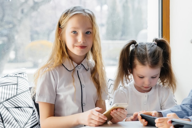 Children sit at a table in a cafe and play mobile phones together. Modern entertainment.